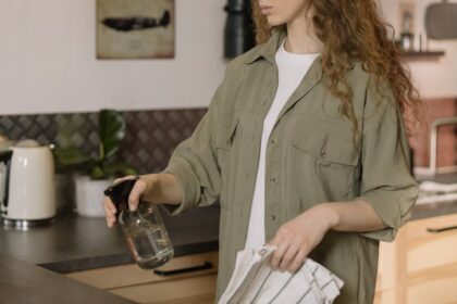 A woman with curly hair cleans a kitchen counter using a spray bottle and cloth.