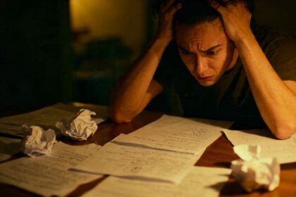 Verwirrte Person sitzt am Tisch, umgeben von Papieren.